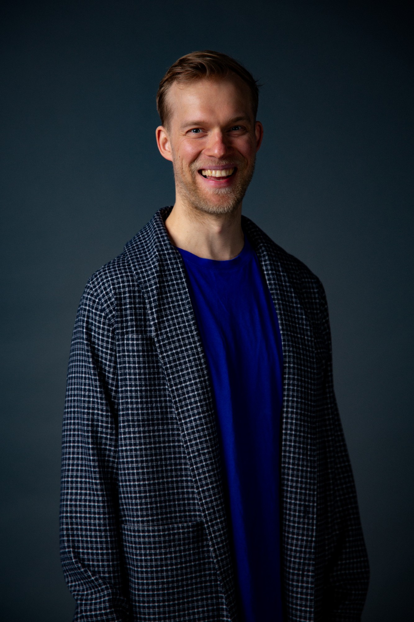 Portrait of Antti Uimonen, smiling, in a checked jacket over a bright blue T-shirt, against a dark blue-grey background.