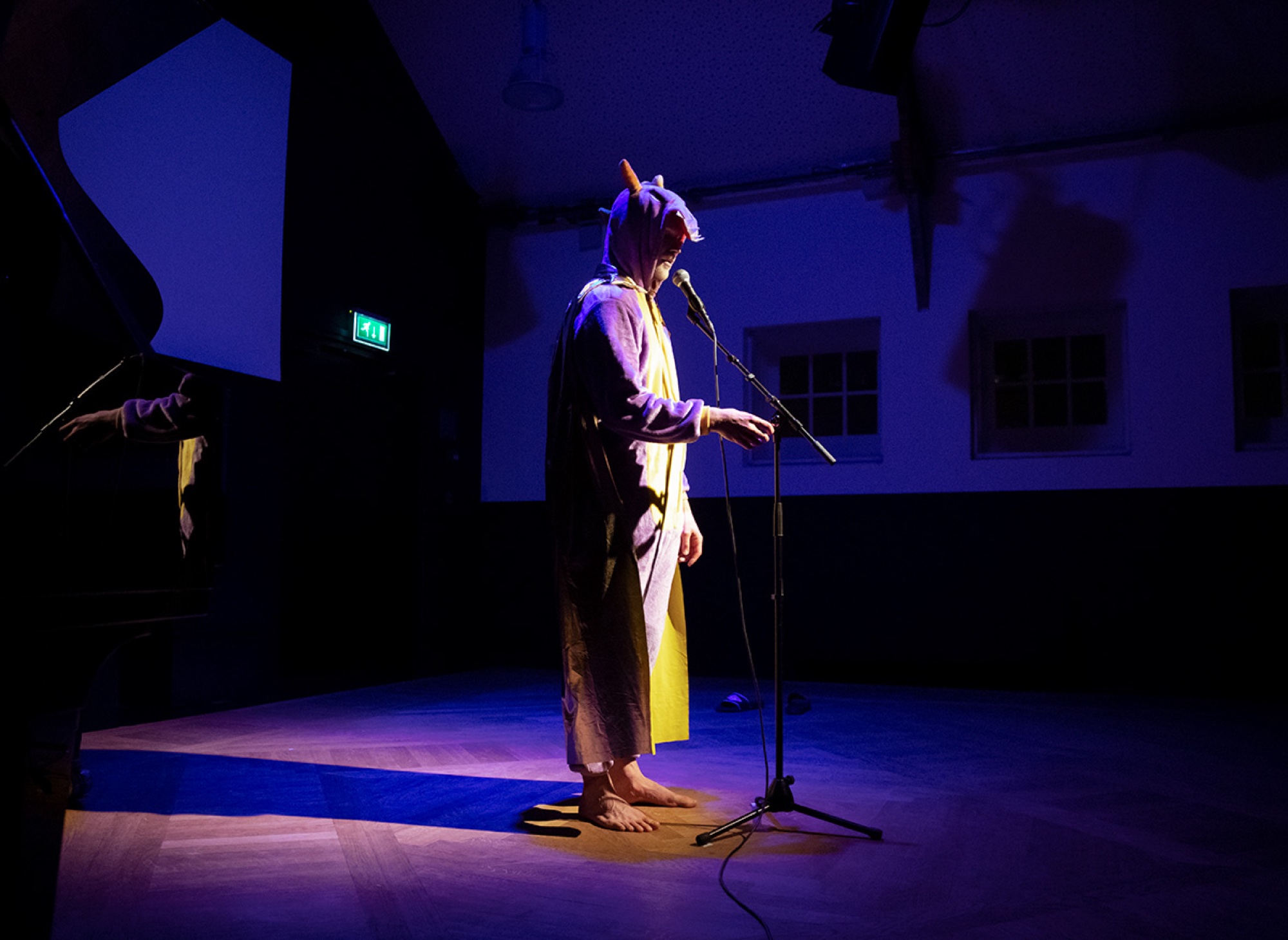 A performer in a purple and yellow creature onesie standing at a microphone in a pool of violet light.