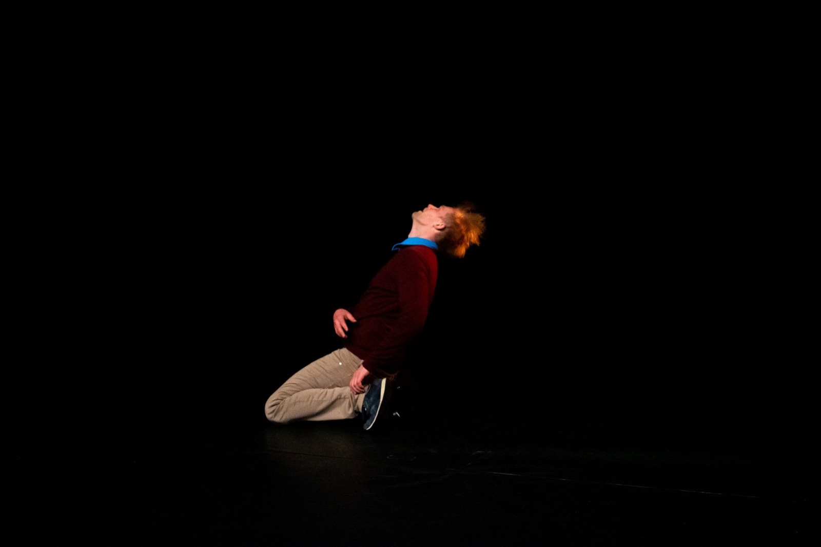 Antti Uimonen kneeling with his head thrown back and hair flying, full body on a dark stage.