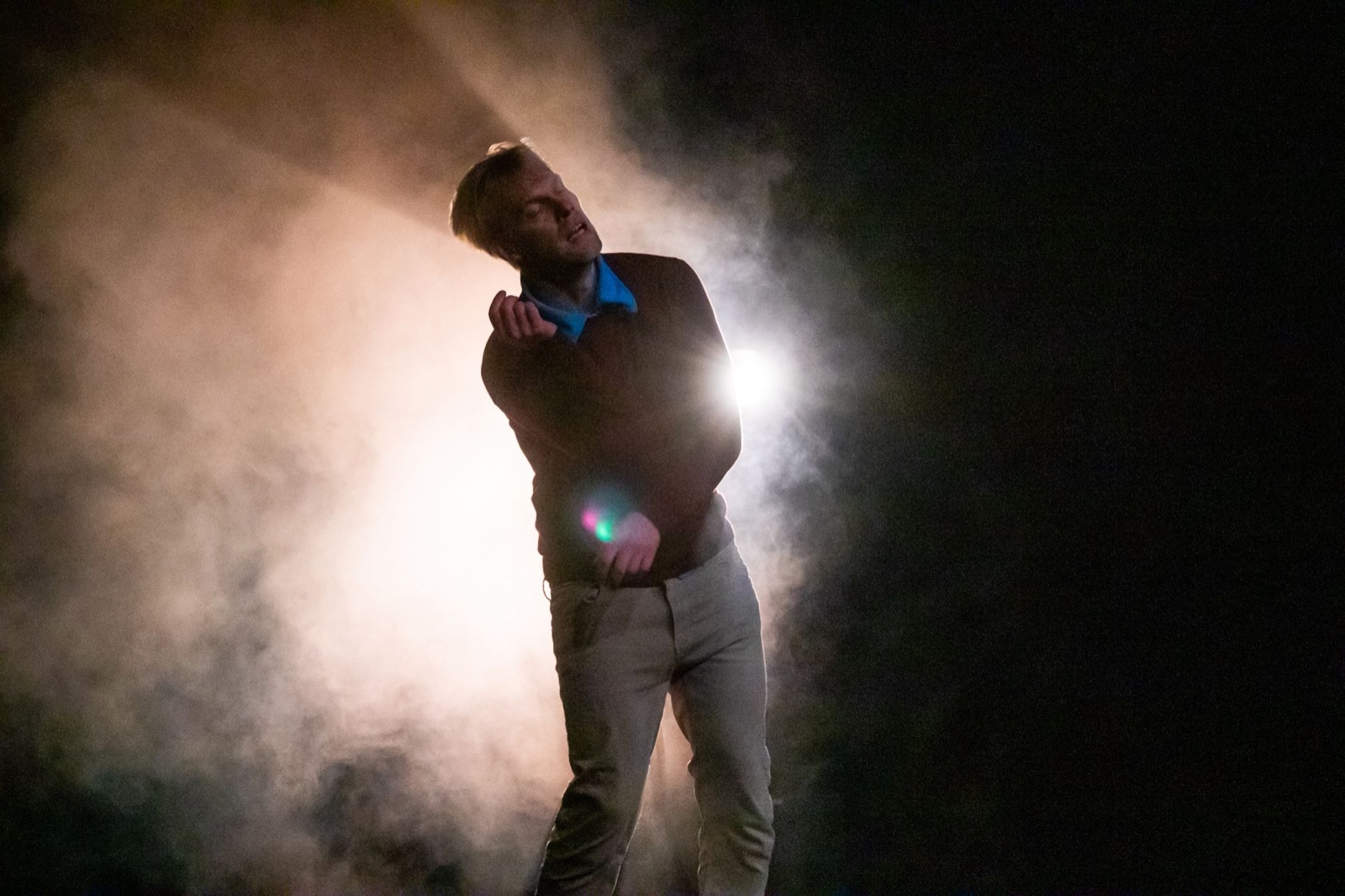 Antti Uimonen leaning forward in hazy stage light, one hand at his collar, looking upward.