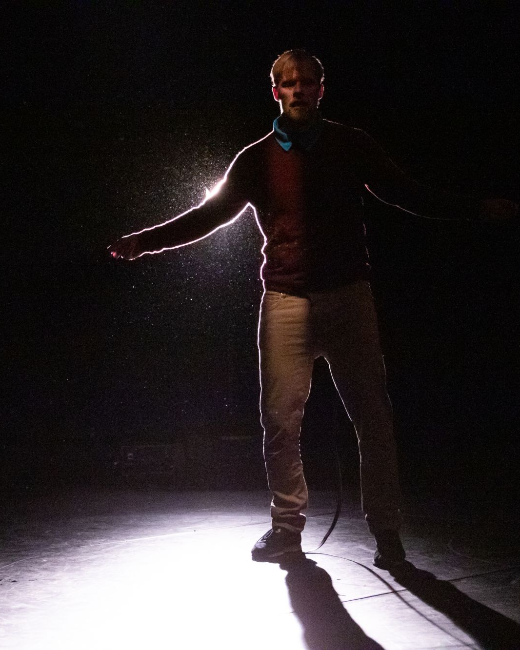 Antti Uimonen standing in a spotlight, one arm outstretched, dust catching the light.