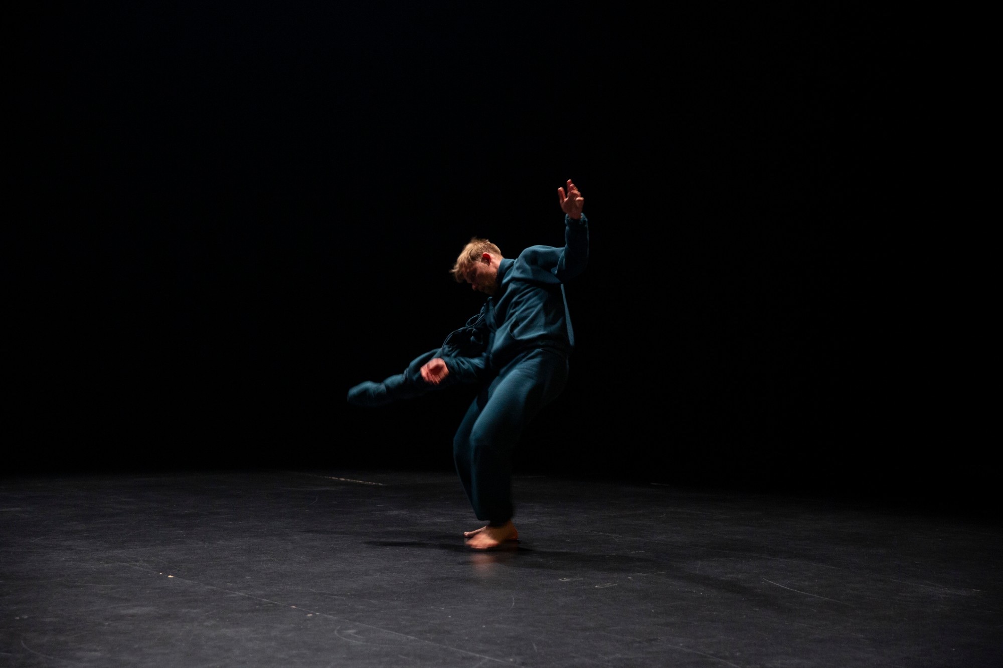 A dancer in a navy tracksuit mid-movement, one arm raised, alone on a dark stage.