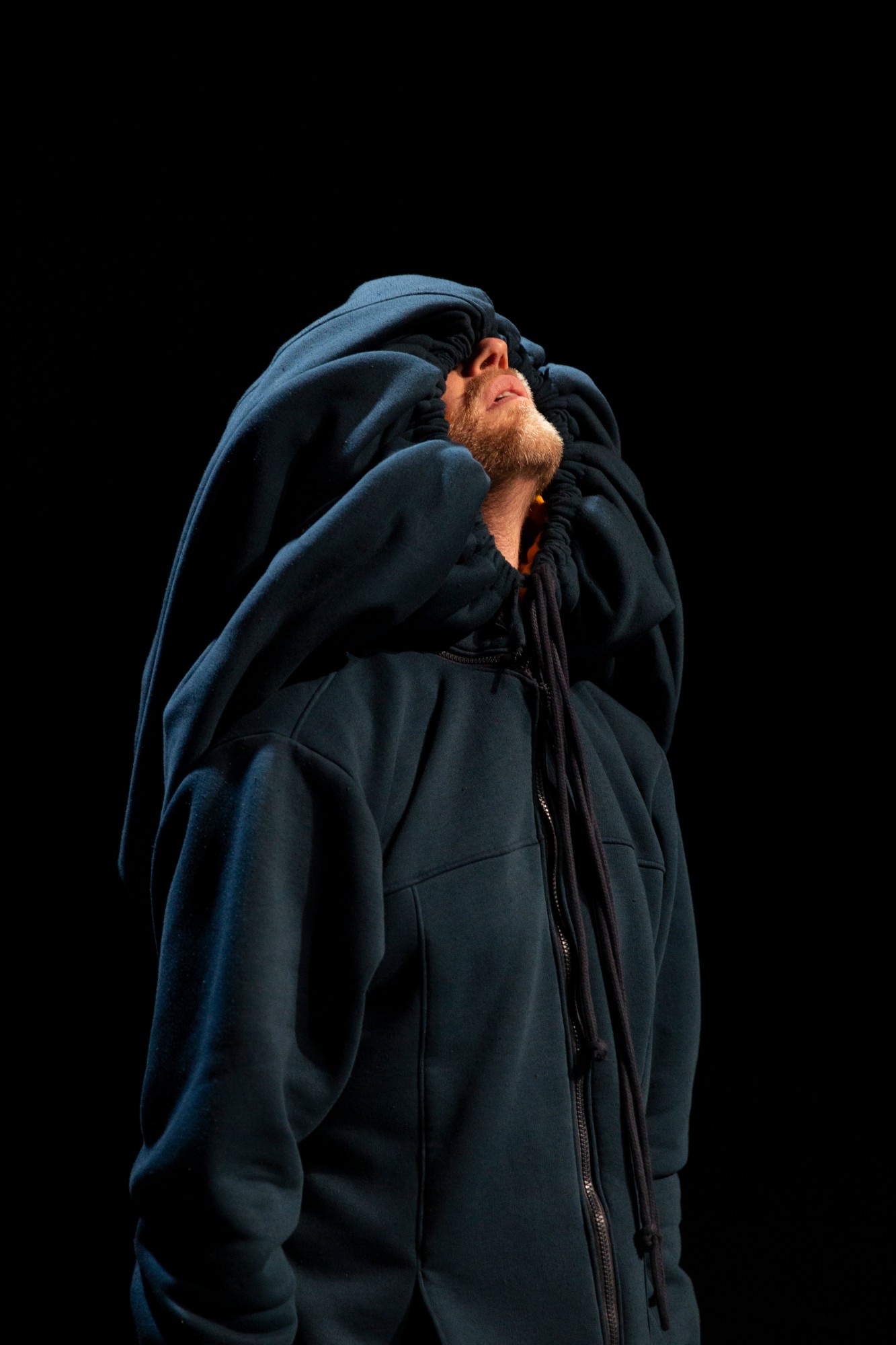 A performer on a black stage in an oversized navy hoodie, the hood pulled up over the face, chin lifted.