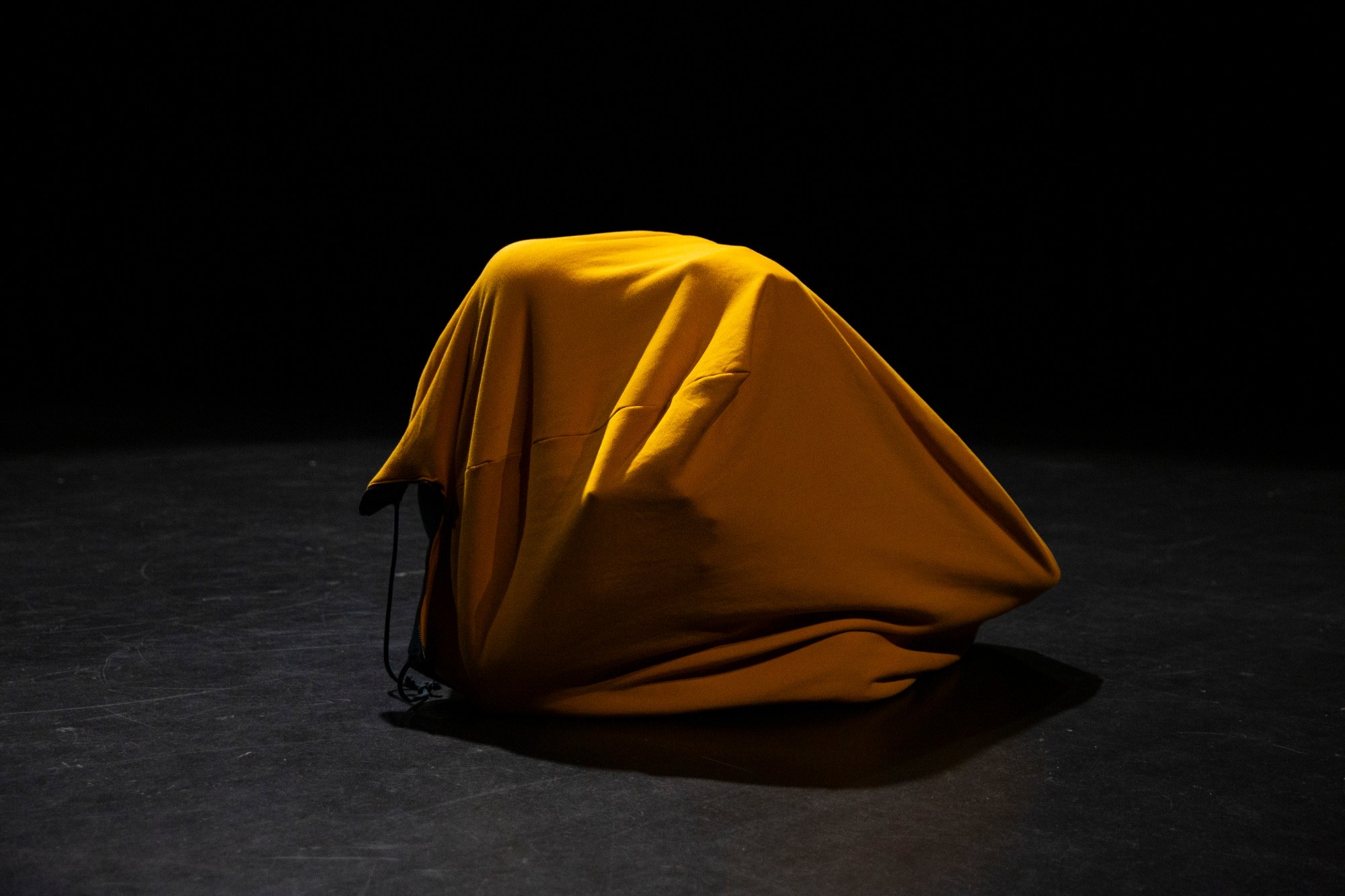 A mustard-yellow cloth draped over a hidden shape on a dark stage.