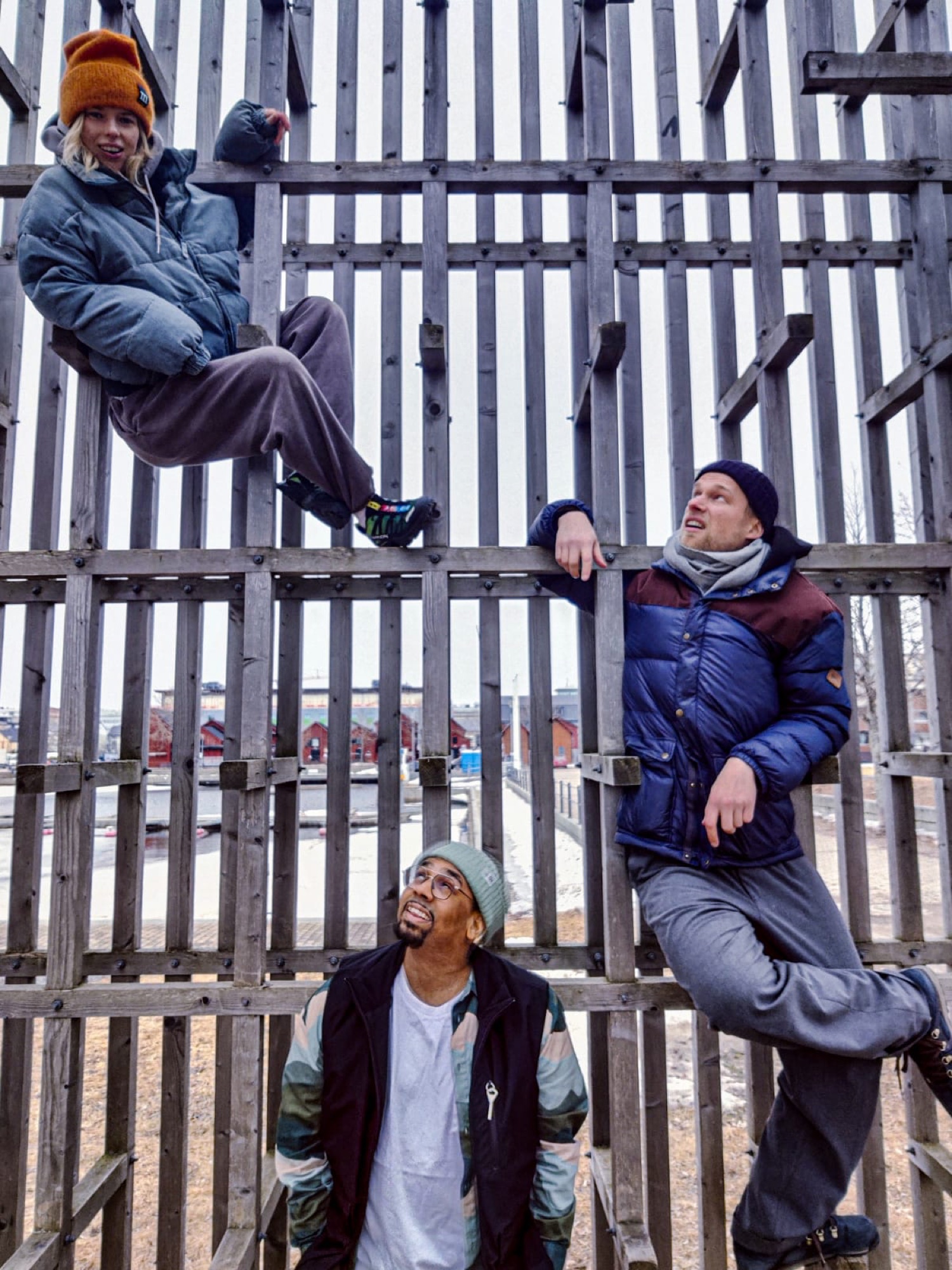 Three people climbing and leaning on a tall wooden slat structure in the snow.